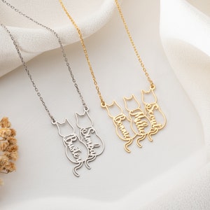 Op de afbeelding: Twee zilveren en gouden kettingen met katten silhouetten en gepersonaliseerde namen. De zilveren ketting heeft de namen "Buster" en "Simba" en de gouden ketting heeft de namen "Bonnie", "Millie" en "Scout".