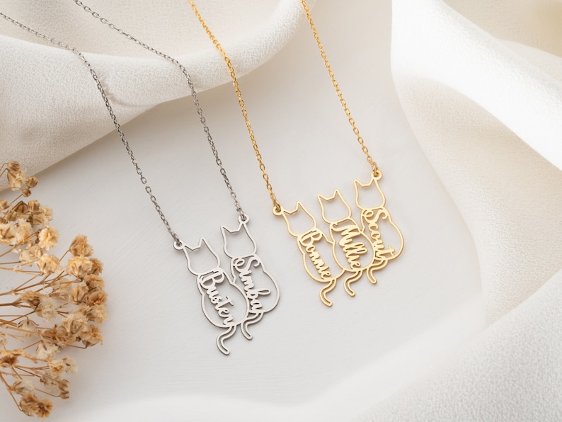 Op de afbeelding: Twee zilveren en gouden kettingen met katten silhouetten en gepersonaliseerde namen. De zilveren ketting heeft de namen "Buster" en "Simba" en de gouden ketting heeft de namen "Bonnie", "Millie" en "Scout".