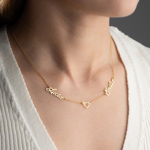 May include: Gold necklace with two names, "Lucia" and "Emilia", written in cursive script, connected by a small heart charm.