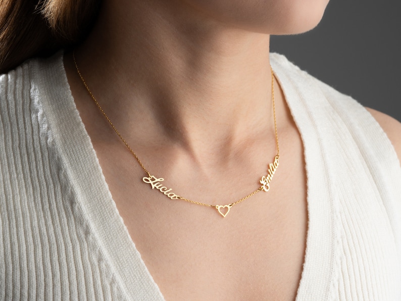 May include: Gold necklace with two names, "Lucia" and "Emilia", written in cursive script, connected by a small heart charm.