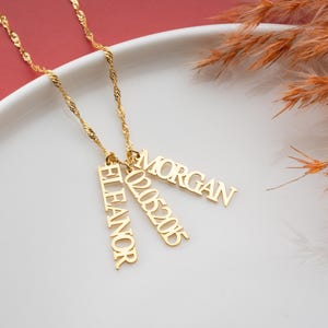 May include: Gold necklace with three pendant charms. The charms are rectangular and feature the names "Eleanor", "Morgan", and the date "02.05.2015".