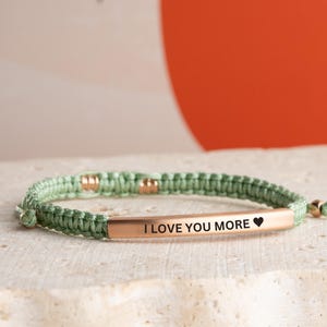May include: A green braided bracelet with a gold bar that says "I love you more" with a small heart symbol.