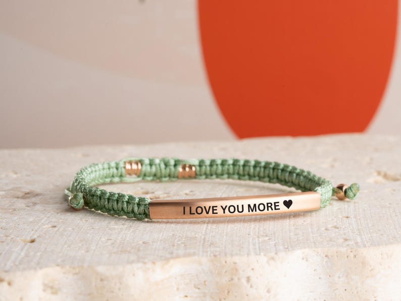May include: A green braided bracelet with a gold bar that says "I love you more" with a small heart symbol.
