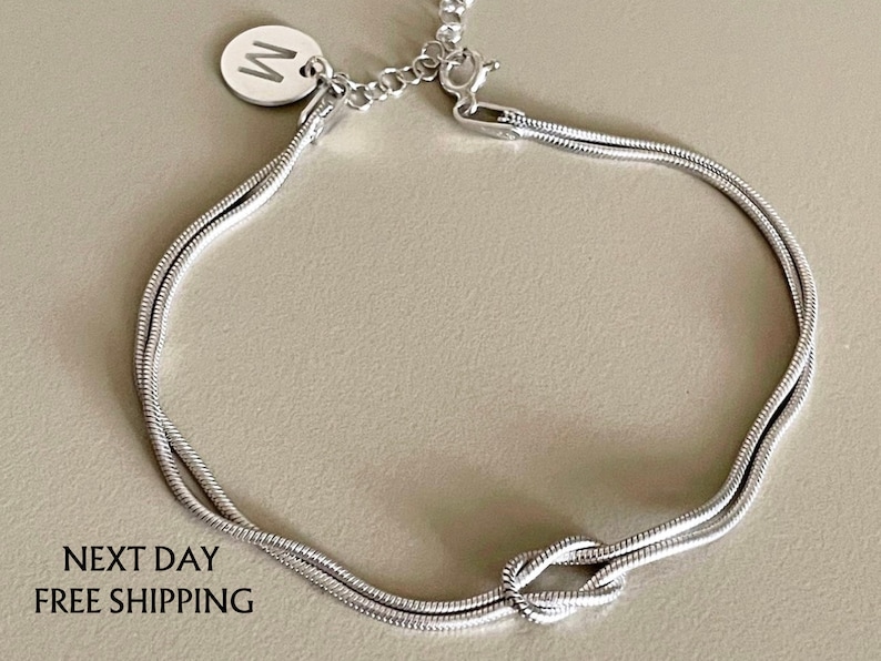 May include: A silver chain bracelet with a knot design and a small silver charm with the letter "W" on it.
