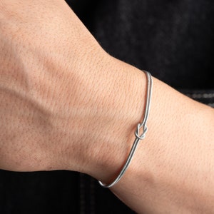 May include: A silver bracelet with a knot design. The bracelet is on a person's wrist.