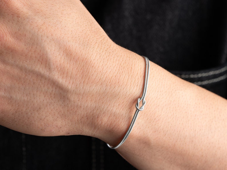 May include: A silver bracelet with a knot design. The bracelet is on a person's wrist.