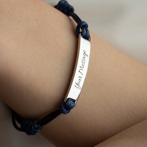 May include: A silver bracelet with a rectangular bar engraved with the words "Your Message" in cursive. The bracelet is attached to a dark blue rope band with knots.