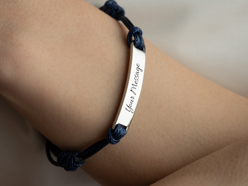 May include: A silver bracelet with a rectangular bar engraved with the words "Your Message" in cursive. The bracelet is attached to a dark blue rope band with knots.