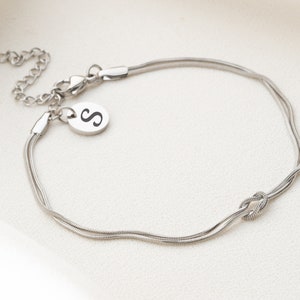 May include: A silver chain bracelet with a knot design and a small charm with the letter "S" engraved on it.
