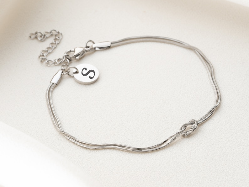 May include: A silver chain bracelet with a knot design and a small charm with the letter "S" engraved on it.