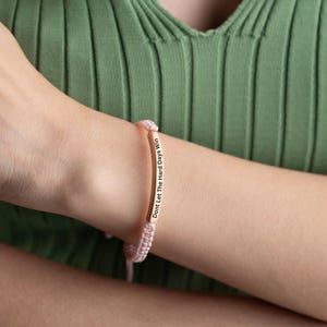 May include: A pink string bracelet with a gold bar charm that reads "Don't Let The Hard Days Win".
