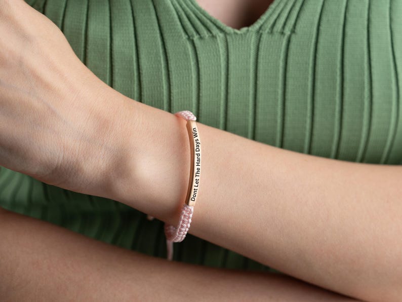 May include: A pink string bracelet with a gold bar charm that reads "Don't Let The Hard Days Win".