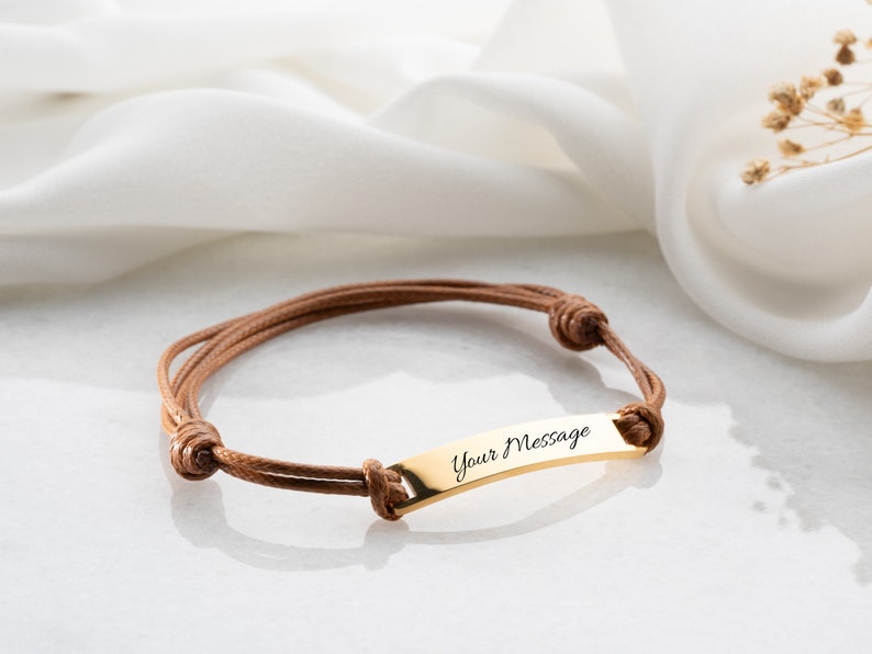 May include: A personalized bracelet featuring a gold-colored rectangular plate with the inscription "Your Message." The plate is attached to a brown cord bracelet. The bracelet is displayed on a white surface with a soft, blurred background.