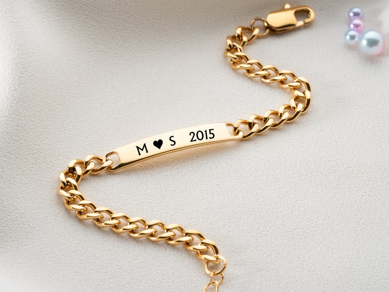 May include: Gold chain bracelet with a personalized bar charm engraved with the letters "M", a heart symbol, "S", and the year "2015".