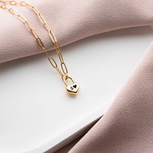 以下が含まれることがあります： ハート型の南京錠のペンダントが付いたゴールドチェーンネックレス。南京錠は黒のエナメル仕上げで、"S"の文字が刻まれています。