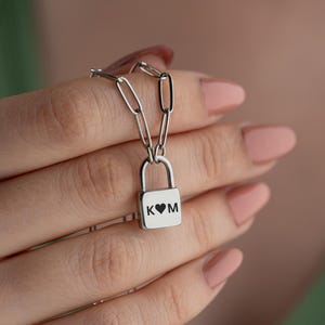 以下が含まれることがあります： ハートとイニシャル「K」と「M」が刻まれた南京錠のペンダントが付いたシルバーチェーンネックレスです。