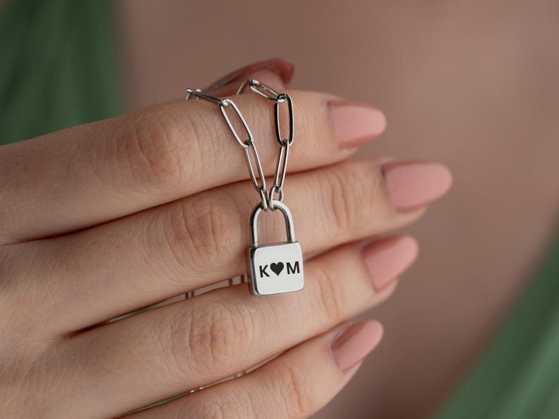 以下が含まれることがあります： ハートとイニシャル「K」と「M」が刻まれた南京錠のペンダントが付いたシルバーチェーンネックレスです。