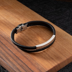 May include: A black leather bracelet with a silver metal accent. The bracelet has a clasp closure.