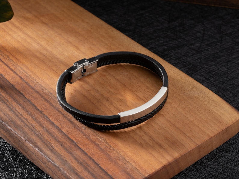 May include: A black leather bracelet with a silver metal accent. The bracelet has a clasp closure.