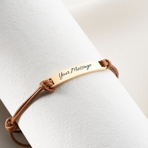 May include: A gold-colored bar bracelet with the text "Your Message" engraved on it. The bracelet is attached to a brown leather cord. The cord is tied with knots on each side of the bar. The bracelet is displayed on a white surface.