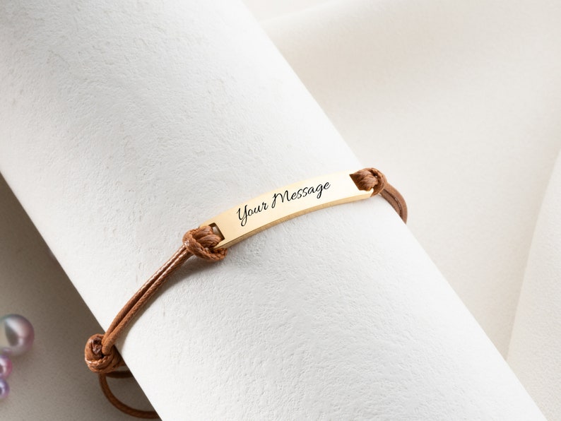 May include: A gold-colored bar bracelet with the text "Your Message" engraved on it. The bracelet is attached to a brown leather cord. The cord is tied with knots on each side of the bar. The bracelet is displayed on a white surface.