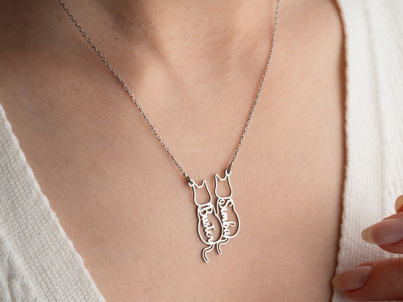 Op de afbeelding: Een zilveren ketting met twee kattenvormige hangers. De hangers zijn gegraveerd met de namen "Simba" en "Buster".