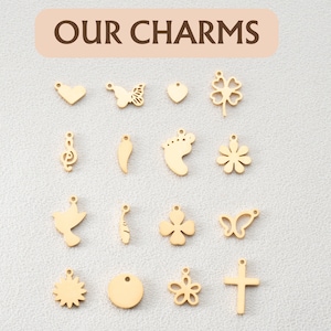 May include: A collection of 15 gold-colored metal charms with various designs, including a heart, butterfly, flower, footprint, musical note, and cross. The charms are arranged in three rows of five on a white background. The text "OUR CHARMS" is written in brown letters on a light pink background at the top of the image.