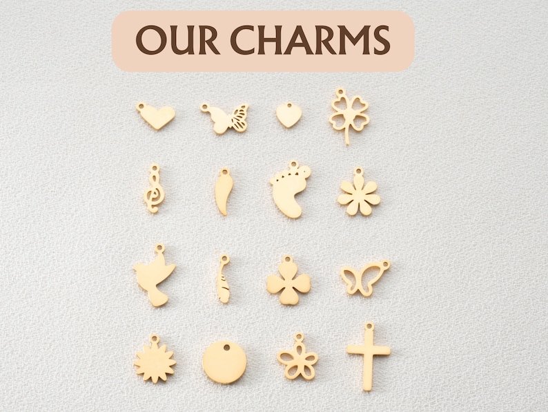 May include: A collection of 15 gold-colored metal charms with various designs, including a heart, butterfly, flower, footprint, musical note, and cross. The charms are arranged in three rows of five on a white background. The text "OUR CHARMS" is written in brown letters on a light pink background at the top of the image.