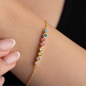May include: A gold-colored bracelet with a delicate chain and a series of colorful gemstones. The gemstones are round and set in a row, featuring various colors like blue, pink, and green. The bracelet is worn on a person's arm.