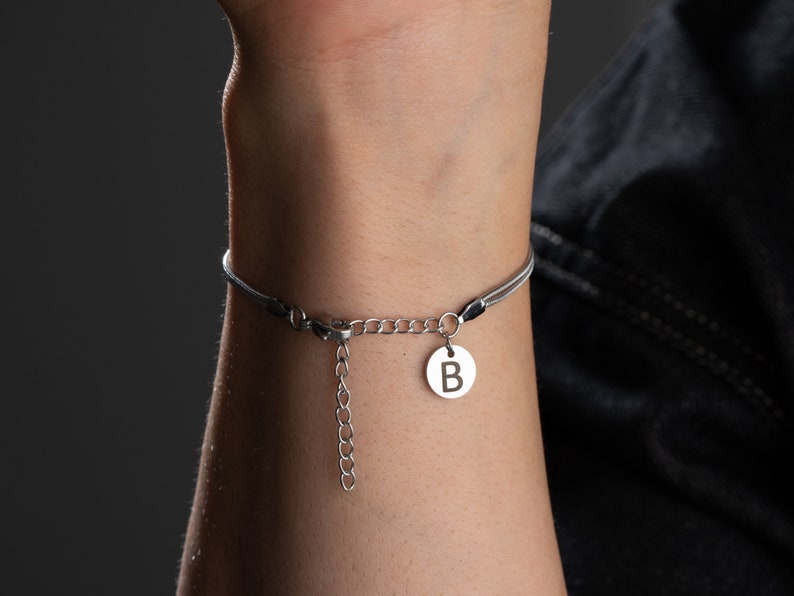 May include: A silver bracelet with a black cord and a silver charm with the letter "B" on it.