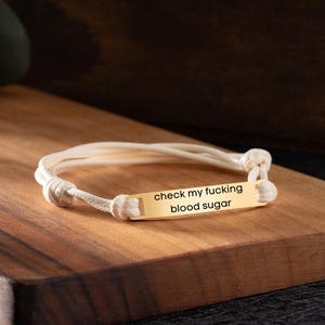 May include: A gold-colored bracelet with the text "check my fucking blood sugar" engraved on a rectangular plate. The bracelet is made of white cord and is knotted at the ends.