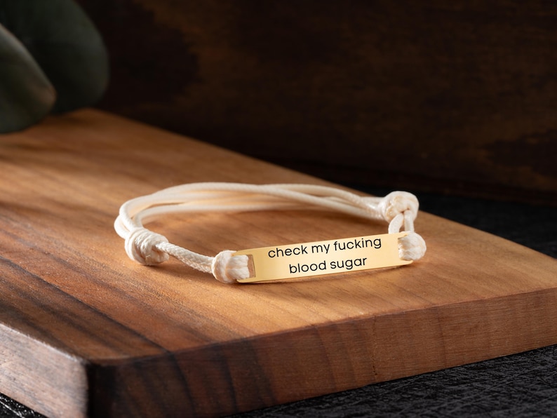May include: A gold-colored bracelet with the text "check my fucking blood sugar" engraved on a rectangular plate. The bracelet is made of white cord and is knotted at the ends.