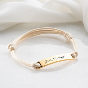May include: A gold-colored bracelet with the text "Your Message" engraved on a rectangular plate. The bracelet features cream-colored rope-like bands and adjustable knots. The bracelet is displayed on a white surface.