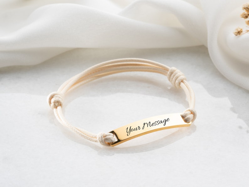 May include: A gold-colored bracelet with the text "Your Message" engraved on a rectangular plate. The bracelet features cream-colored rope-like bands and adjustable knots. The bracelet is displayed on a white surface.