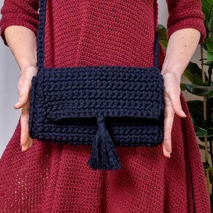 May include: A navy blue crochet crossbody bag with a flap closure and a tassel. The bag is handmade with a thick yarn, and has a long strap. The bag is held in front of a person wearing a red dress.