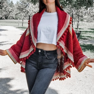 May include: A red and pink crocheted shawl with a pom-pom trim. The shawl has a striped design with shades of pink, cream, and gold. The sleeves are wide and the shawl is worn over a white top and dark jeans.