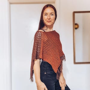 May include: A rust-colored, open-weave crochet poncho with fringe. The poncho has a loose, flowing design and is made of a soft, textured yarn. The garment is a stylish accessory for cooler weather.