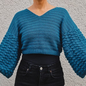 Könnte beinhalten: Ein türkisfarbener Häkelpullover mit V-Ausschnitt und strukturierten Ärmeln.