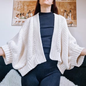 Pode incluir: Um cardigan de malha de cor creme com um estilo solto de abertura frontal e mangas compridas. O cardigan é feito de um fio macio e texturizado e tem um caimento relaxado.