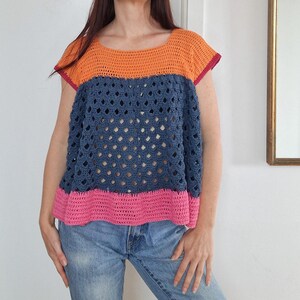 May include: A handmade crochet top in orange, blue, and pink. The top features a square neckline and short sleeves with a magenta trim. The body of the top has an open, lacy design. The bottom hem is a bright pink.
