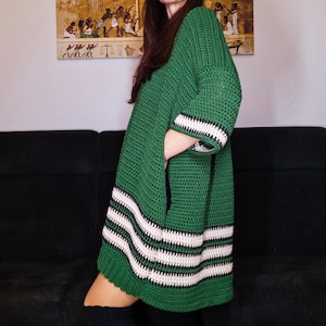 以下が含まれることがあります： 裾に白と黒のストライプが入った緑色のクロシェ編みドレス。ゆったりとしたシルエットで、スカートは少し広がっています。