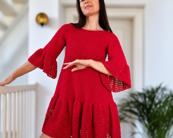 Pode incluir: Um vestido de crochê vermelho vibrante com decote redondo, mangas compridas com punhos largos em sino e uma bainha com babados. A peça é confeccionada com um padrão de ponto texturizado e aberto, criando um visual consistente em toda a peça.