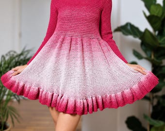 Pode incluir: Um vestido de crochê de manga comprida com uma parte superior em fúcsia vibrante que transita para uma saia em degradê rosa claro. A peça tem decote redondo, corpete justo e uma saia rodada e plissada com uma bainha de babados na base.