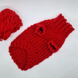 May include: A handmade red crochet dog sweater with armholes and a ribbed neckline and hem. A ball of red yarn is next to the sweater. The sweater is made of a thick, textured yarn.