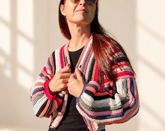 Può includere: Un cardigan a maniche lunghe con chiusura a bottoni, caratterizzato da un motivo all'uncinetto multicolore con strisce orizzontali nei toni del rosso, rosa, nero, bianco, blu e beige. Il capo presenta uno scollo a V, polsini a coste e bottoni rotondi chiari.