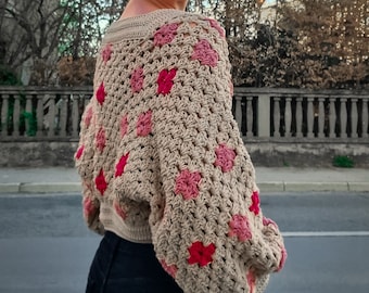 Pode incluir: Um suéter de crochê bege com detalhes de flores rosa. O suéter tem um caimento solto e mangas compridas.