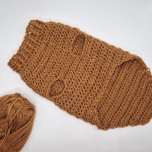 May include: A brown, handmade crochet dog sweater with two armholes. The sweater is made of a thick yarn and has a textured appearance. A ball of matching brown yarn is next to the sweater. The sweater is on a white surface.