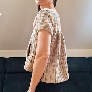 May include: A beige crochet top with short sleeves and a loose fit. The top has a unique design with a cutout on the side.