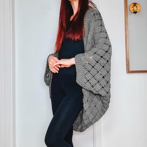 May include: A gray, open-weave crochet cardigan with wide sleeves. The cardigan has a diamond pattern and is worn over a black top and leggings. The sleeves have button details. The cardigan is a comfortable, stylish layering piece.