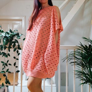 Puede incluir: Un vestido de crochet coral con un ajuste holgado y hombros descubiertos. El vestido tiene un escote cuadrado y un patrón texturizado.